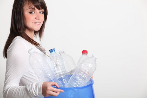 A young woman with shoulder-length brown hair and a light complexion is smiling and standing against a plain white background, holding a blue plastic bin filled with multiple empty plastic bottles of various sizes, including clear bottles with blue and red caps. The bottles appear to be made of PET plastic and are loosely arranged inside the bin, some leaning against the sides. She is wearing a white long-sleeved top, and her left hand is gripping the side of the bin while her right hand supports its base. The environment appears to be indoors, with no visible environmental or outdoor elements, and the lighting is bright and even, emphasizing the clear plastic bottles and her cheerful expression. This image subtly relates to waste collection or recycling activities, aligning with rubbish removal services such as those offered by Commercial Waste Removal Croydon in the local Croydon area, promoting responsible disposal of plastic waste.