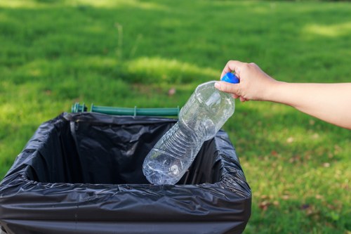 A person's hand is shown holding a semi-transparent plastic water bottle with a blue cap, in the process of discarding it into a black plastic rubbish bin lined with a black bag. The bin is situated outdoors on a paved surface, with a background of well-maintained green grass and blurred shrubbery, indicating a tidy outdoor environment. The scene illustrates typical waste disposal activity, relevant to local rubbish removal services, such as those offered by Commercial Waste Removal Croydon, operating within the Croydon area. The image emphasizes the importance of proper waste segregation and disposal, with the individual's task highlighting routine refuse collection practices in residential or commercial settings.