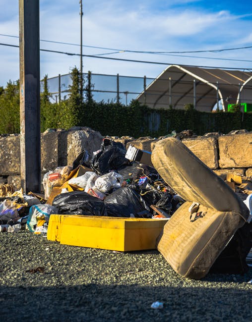 A close-up view of a grey and blue work shoe with yellow accents, partially buried among a large pile of dark, irregularly shaped rocks and discarded cigarette butts scattered across the surface. The 