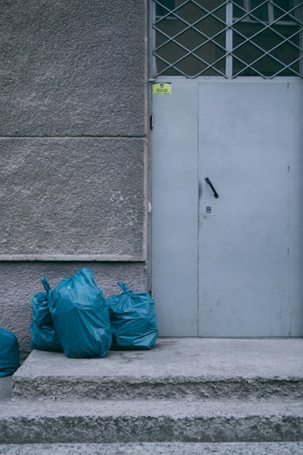 A pile of discarded household items and debris positioned on a paved outdoor surface, including a wooden slatted chair frame leaning against a wall, and various broken or damaged objects such as a whi