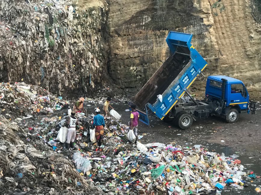 A scene of waste removal featuring a blue skip truck dumping a large quantity of mixed household and commercial rubbish into a waste disposal site. The truck, positioned on the right, has its hydrauli